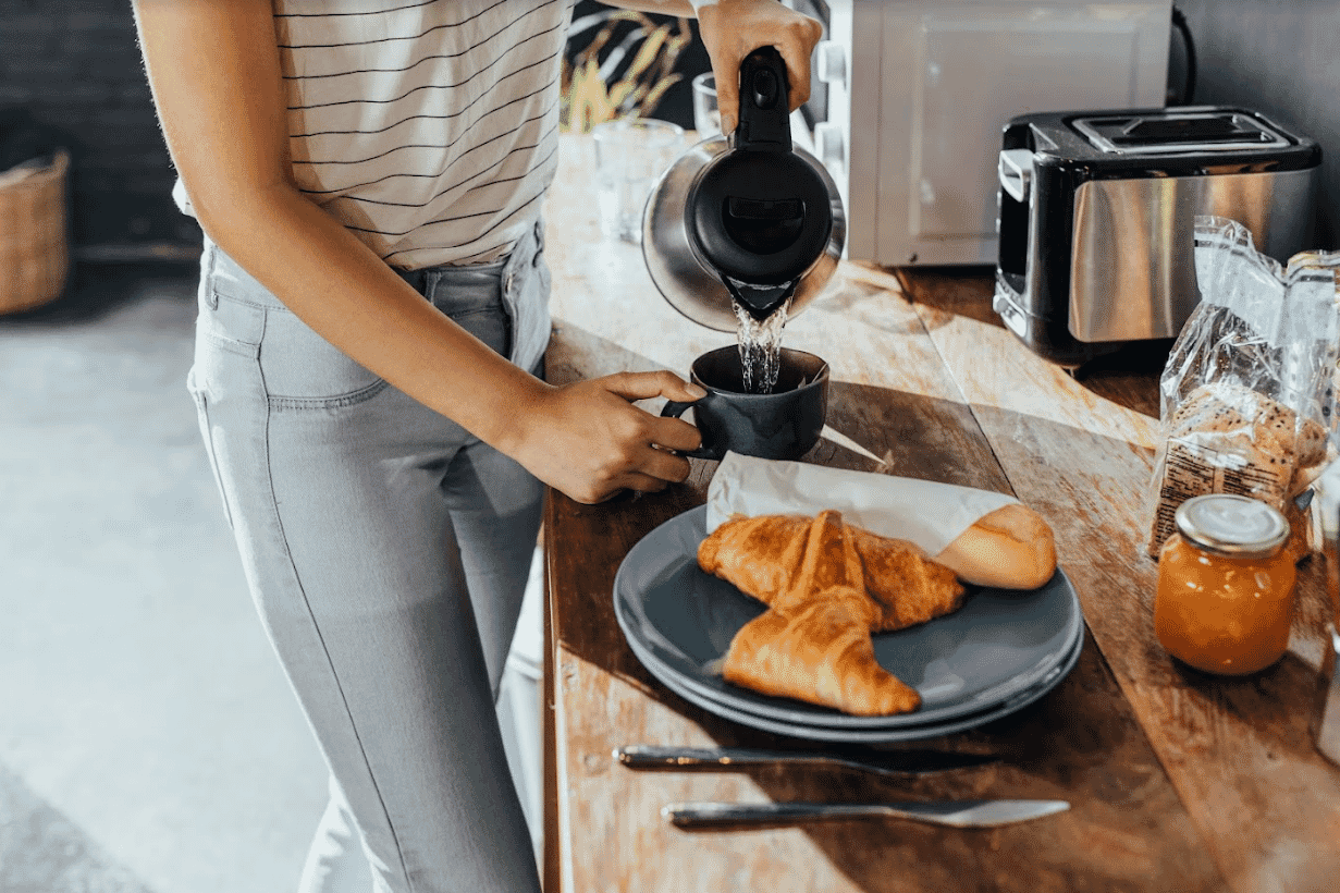 Woman pours hot water from a kettle into a cup with a tea bag at her kitchen bench 