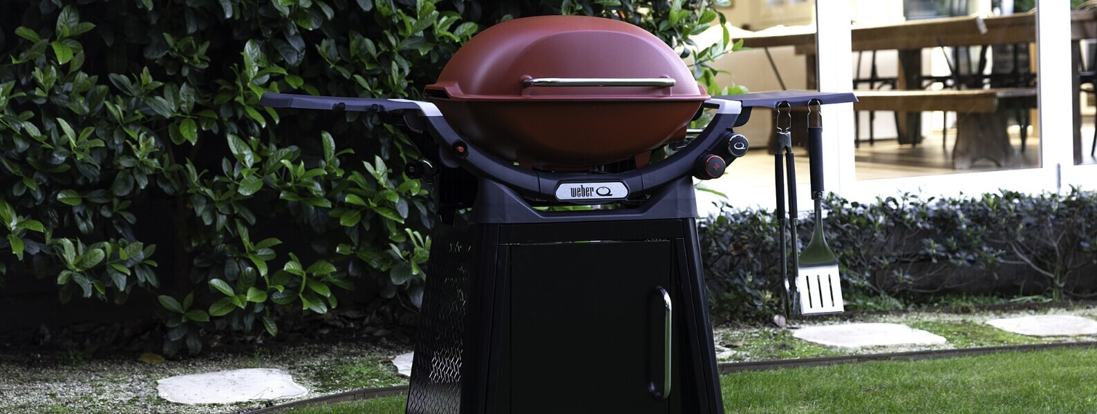 A black Weber Family Q BBQ stands next to an outdoor kitchen in a backyard