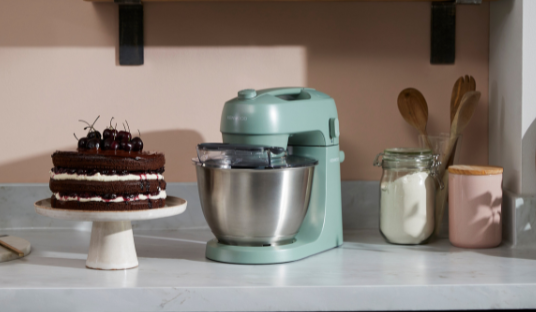 Image of the Kenwood Go Eucalyptus Green Stand Mixer on a bench next to a baked cake.