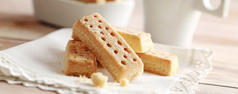 A stack of rectangular shaped shortbread biscuits on a white napkin with a mug and more shortbread in a bowl