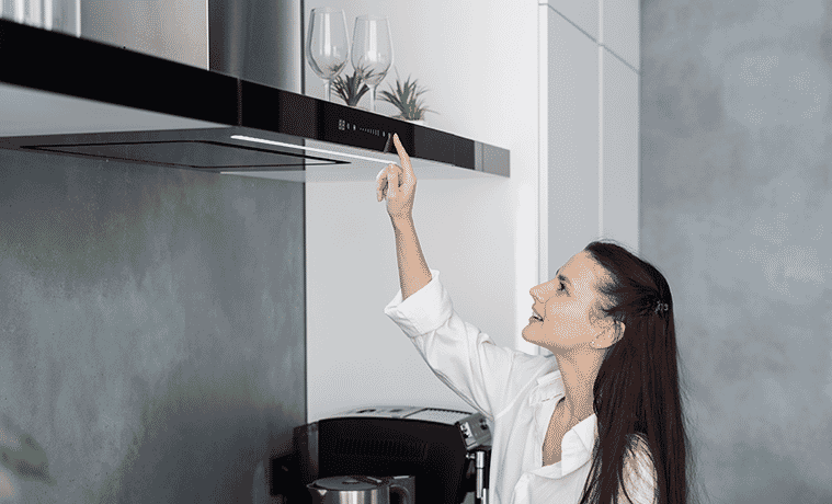 A woman reaches up to flick a switch on the sleek rangehood above the cooktop in her contemporary kitchen