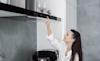 A woman reaches up to flick a switch on the sleek rangehood above the cooktop in her contemporary kitchen
