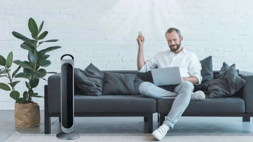 Man sitting on a dark grey couch on his laptop with a tower fan in front of the couch
