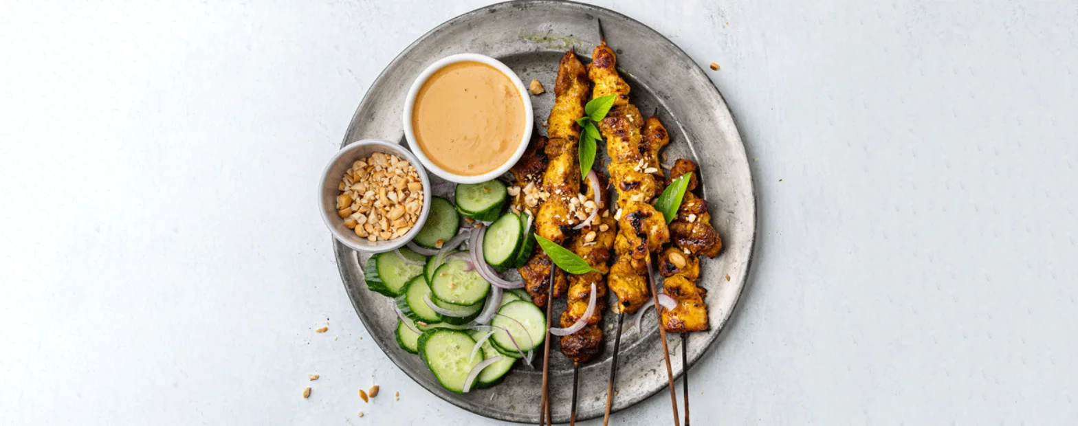 Chicken satay skewers on a pewter plate with peanut sauce, crushed peanuts, and a cucumber and red onion salad.