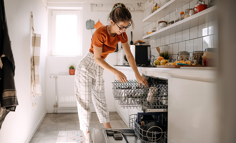 A young woman loads glasses into the top rack of a builtin dishwasher in her white galleystyle kitchen