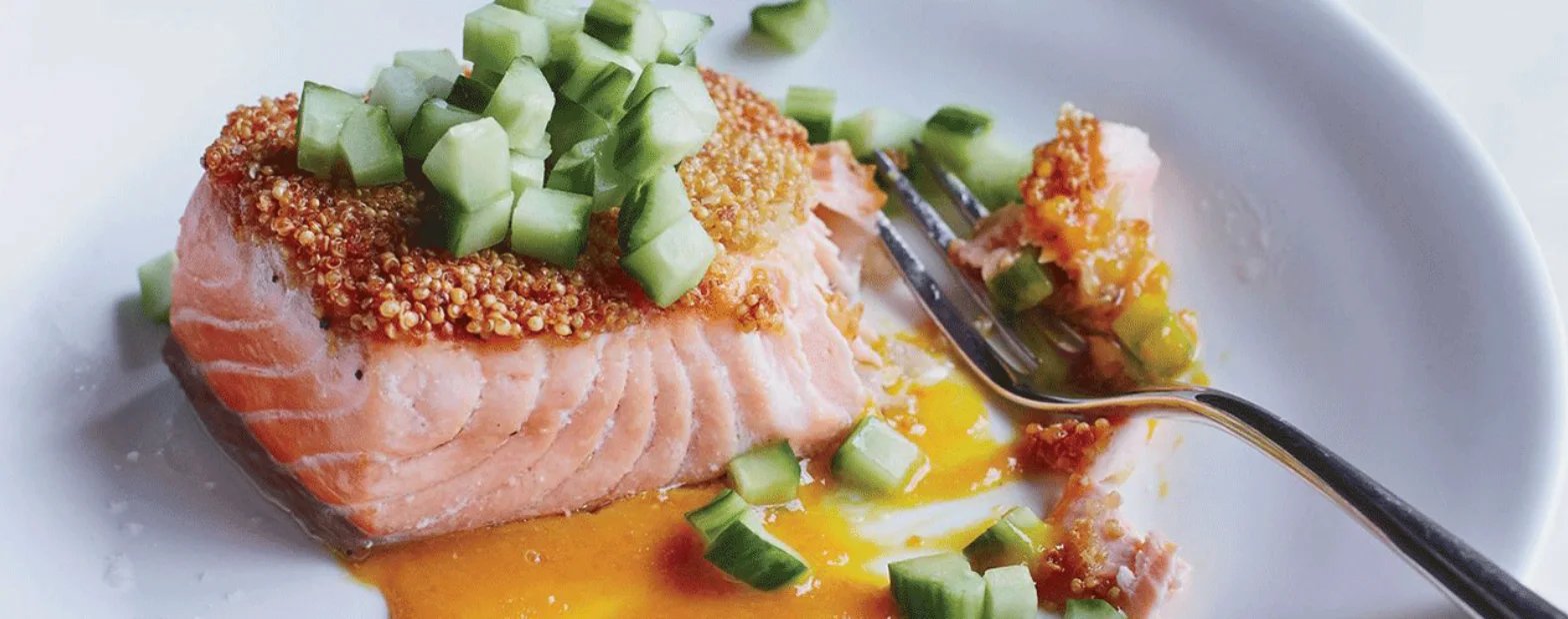 Quinoa-crusted salmon fillet topped with diced cucumber on a white plate with an orange sauce.