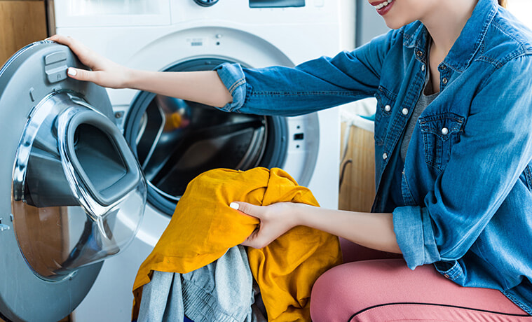 A smiling young woman removes freshly dried clothes from the heat pump dryer in her laundry