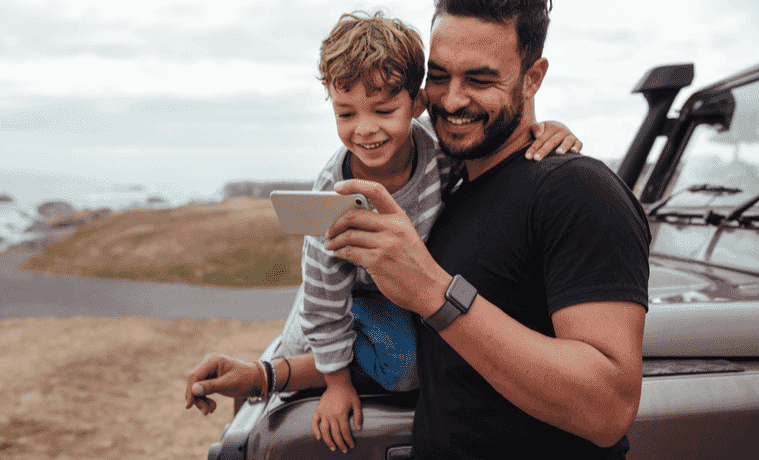 Dad and young son standing next to their 4WD near the coast are both looking at a mobile phone and smiling