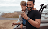 Dad and young son standing next to their 4WD near the coast are both looking at a mobile phone and smiling
