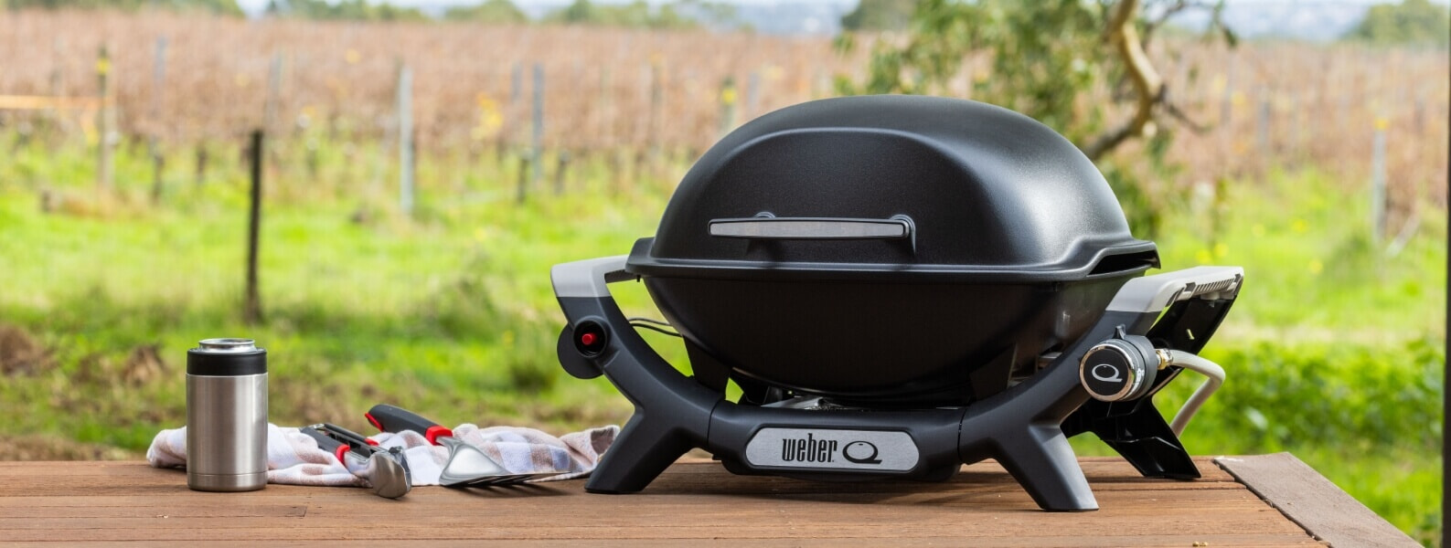 A black Weber Baby Q BBQ sits on a picnic table at a vineyard