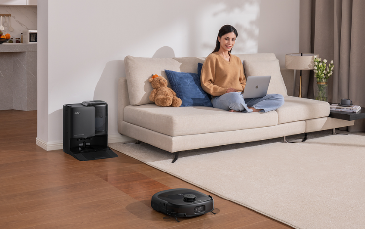 eufy robot vacuum cleaning a living room floor while a woman relaxes on the couch with a laptop
