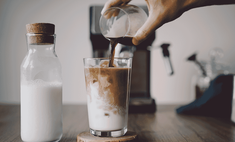 Closeup of a man pouring espresso into a glass of milk with ice next to a bottle of milk with a coffee machine in the background