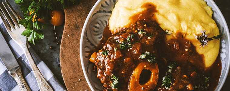 Osso buco beef stew with creamy polenta in a decorative plate on a timber board with antique cutlery and a navy and white checked napkin 