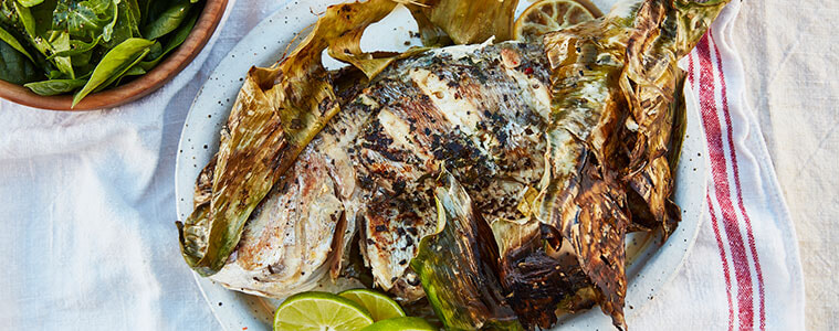 Whole grilled fish on white plate with lime slices next to a bowl of spinach leaves all sitting on a white teatowel