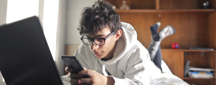 A tween boy lays on his bed and uses his phone in front of his laptop