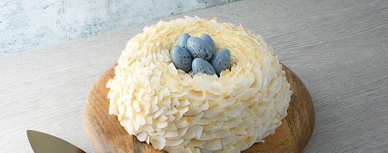 Easter Nest Cake covered in coconut flakes with blue eggs on the top with the cake on a wooden board with a brass cake server