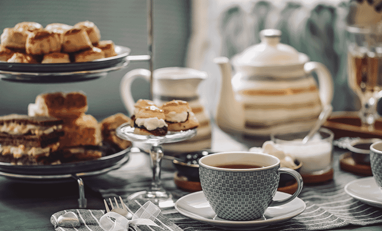A table laid for afternoon tea with a teapot grey china cup and saucer cake stand topped with scones with jam and cream and assorted cakes on a tiered cake stand