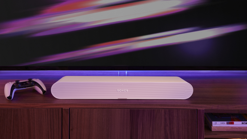 Close up of the Sonos Ray soundbar in white sitting on a wooden TV cabinet next to a gaming console