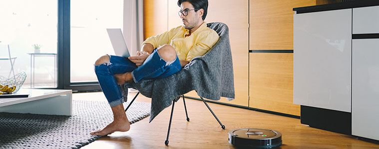 Man at home working on his laptop while a robot vacuum cleaner cleans the floor