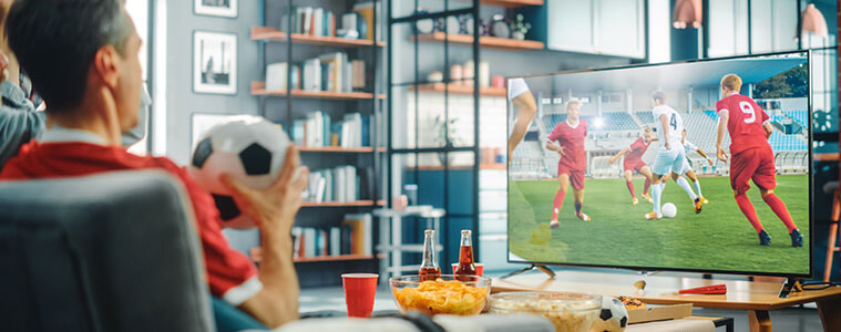 A man sits in his armchair soccer ball in hand and drinks and crisps on the coffee table as he watches soccer on TV