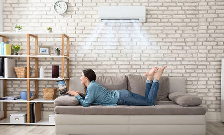 A woman lays on her sofa and uses her laptop while her split system air conditioner keeps her cool