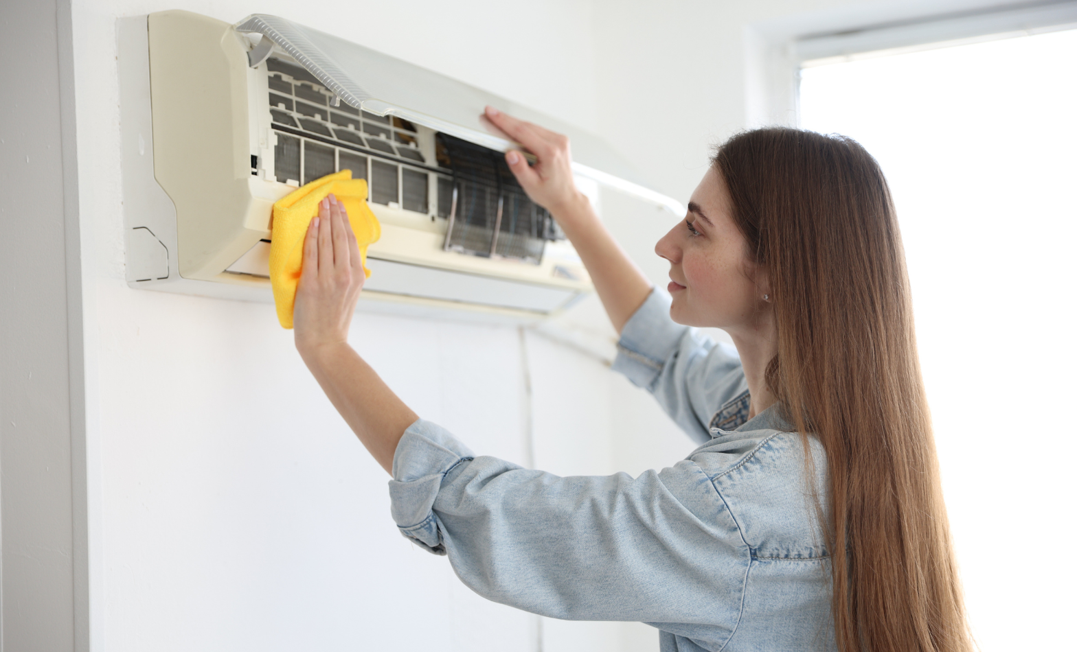 Woman using a yellow microfibre cloth to clean the open front panel of a wall-mounted split system air conditioner