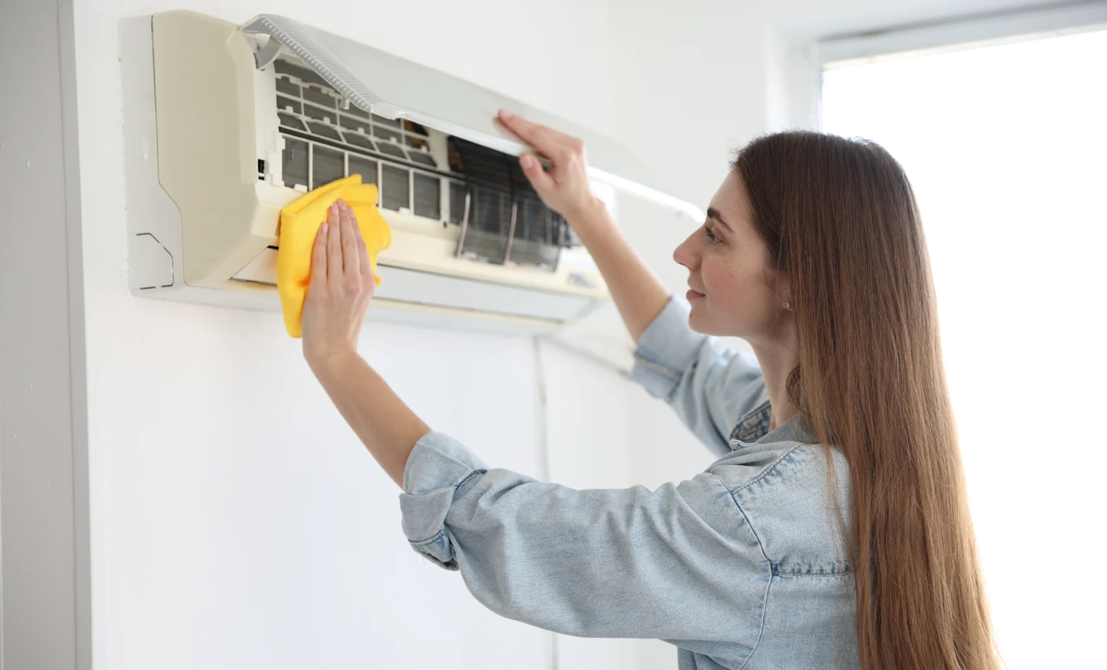 Woman using a yellow microfibre cloth to clean the open front panel of a wall-mounted split system air conditioner