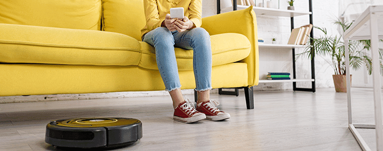 Young girl with a smartphone on a sofa smiling and looking at a robotic vacuum cleaner on the living room floor