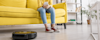 Young girl with a smartphone on a sofa smiling and looking at a robotic vacuum cleaner on the living room floor