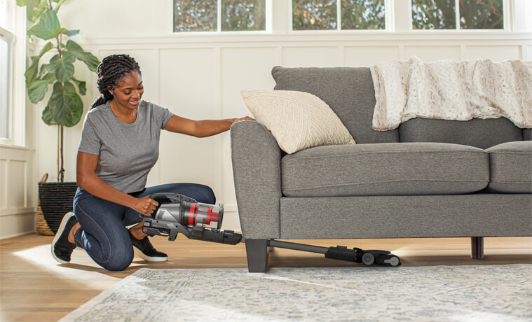 A woman vacuums under her couch with a Hoover stick vacuum