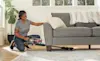 A woman vacuums under her couch with a Hoover stick vacuum
