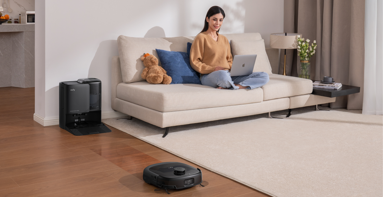 eufy robot vacuum cleaning a living room floor while a woman relaxes on the couch with a laptop