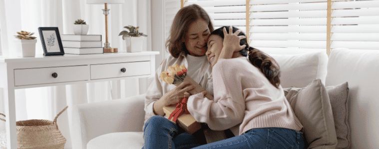 Older woman holds a  gift box and flowers in her hand while hugging a younger woman as they sit together on a couch