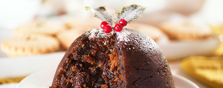 A Christmas pudding on a white plate with a dusting of icing sugar and holly decoration on top