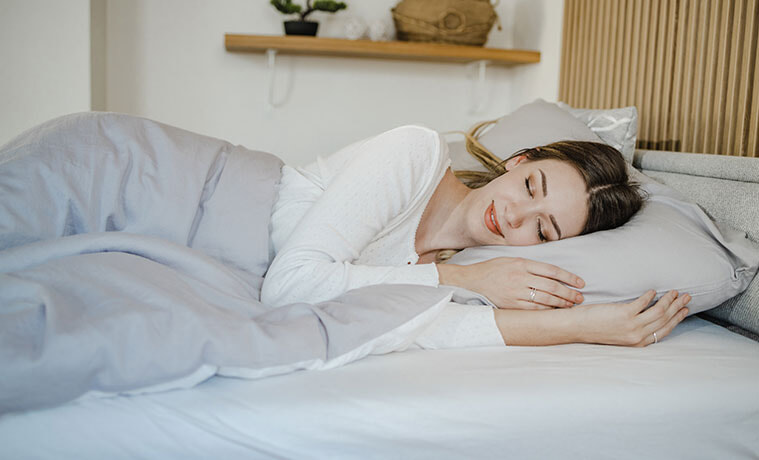 A young woman smiling in bed with eyes closed under a cosy grey doona