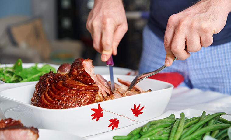 Closeup of a man using a knife and fork to carve a glazed ham at Christmas 