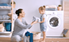 A mother hands her young daughter some freshly dried clothes from a basket in their laundry room