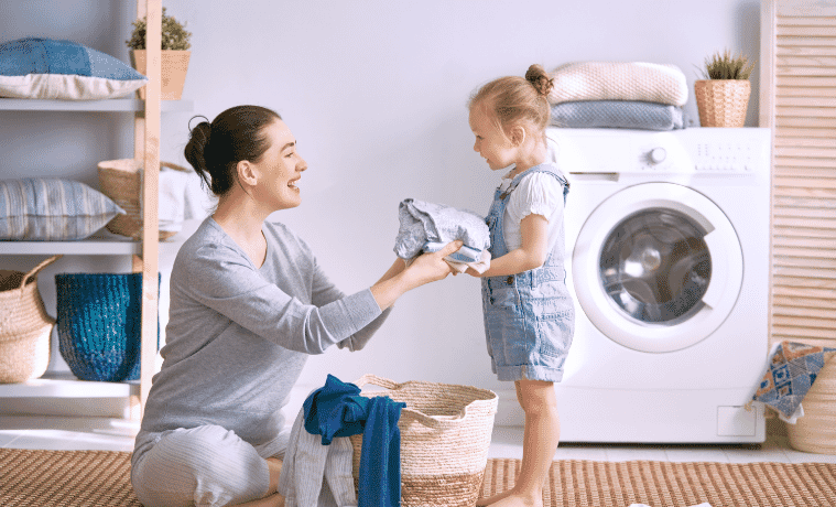 A mother hands her young daughter some freshly dried clothes from a basket in their laundry room