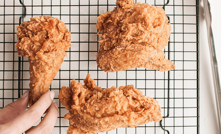 Closeup of a hand reaching for a fried chicken drumstick on a wire rack with two other pieces of fried chicken