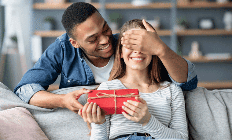 A man covers the eyes of a woman sitting on a couch as he leans over from behind the couch and hands her a gift
