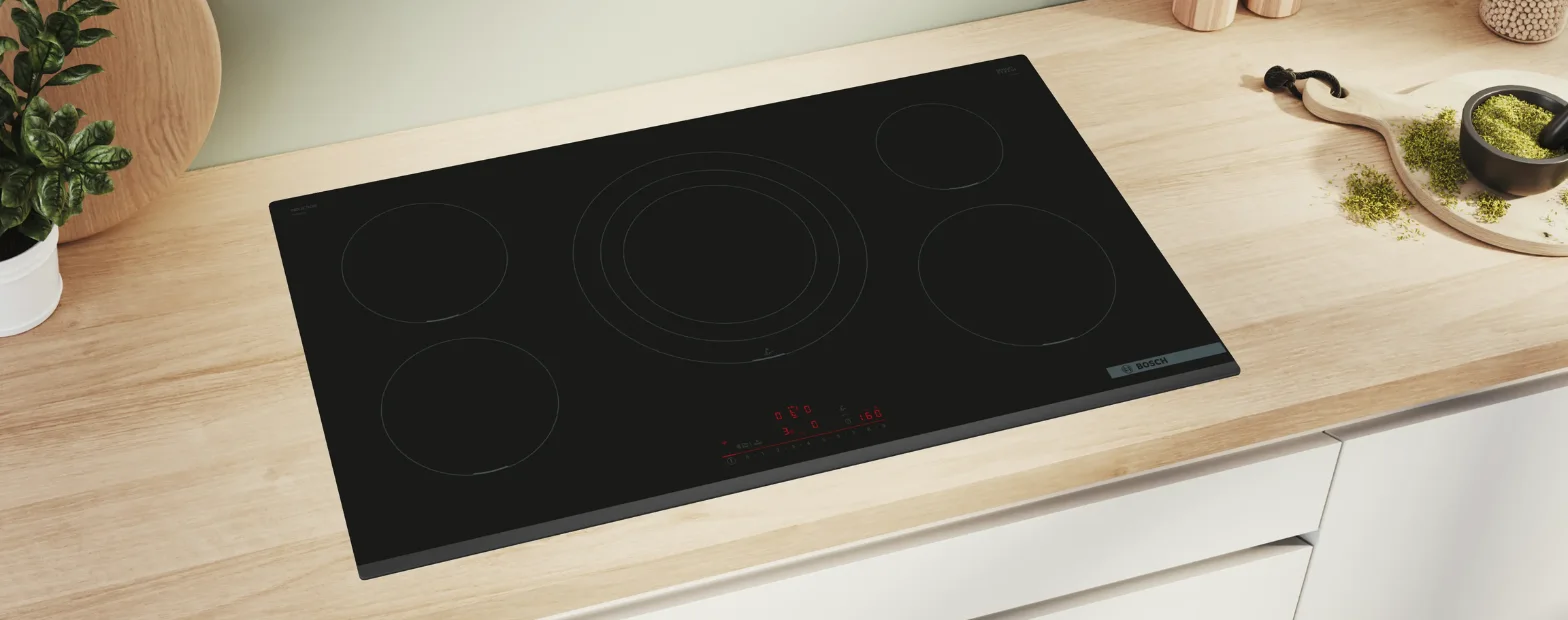 Bosch black induction cooktop with five cooking zones and red digital touch controls, installed in a light timber benchtop with a basil plant and herbs nearby.