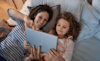A mother and daughter lie on a bed at home and watch a TV show on a tablet