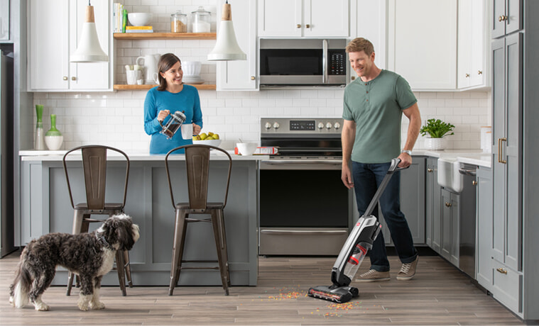 A man vacuums his kitchen while his wife and dog watch