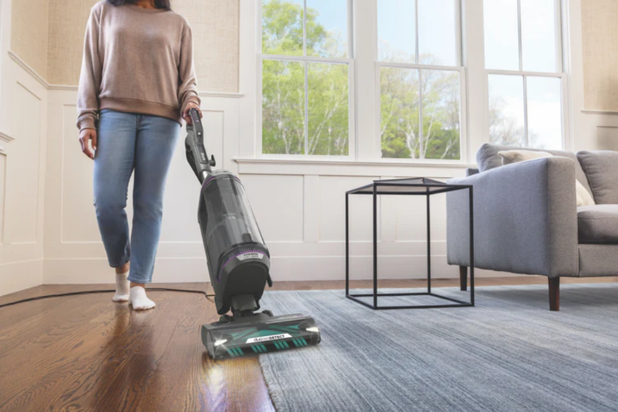 Woman using Shark Upright Vac to vacuum her living room;. 