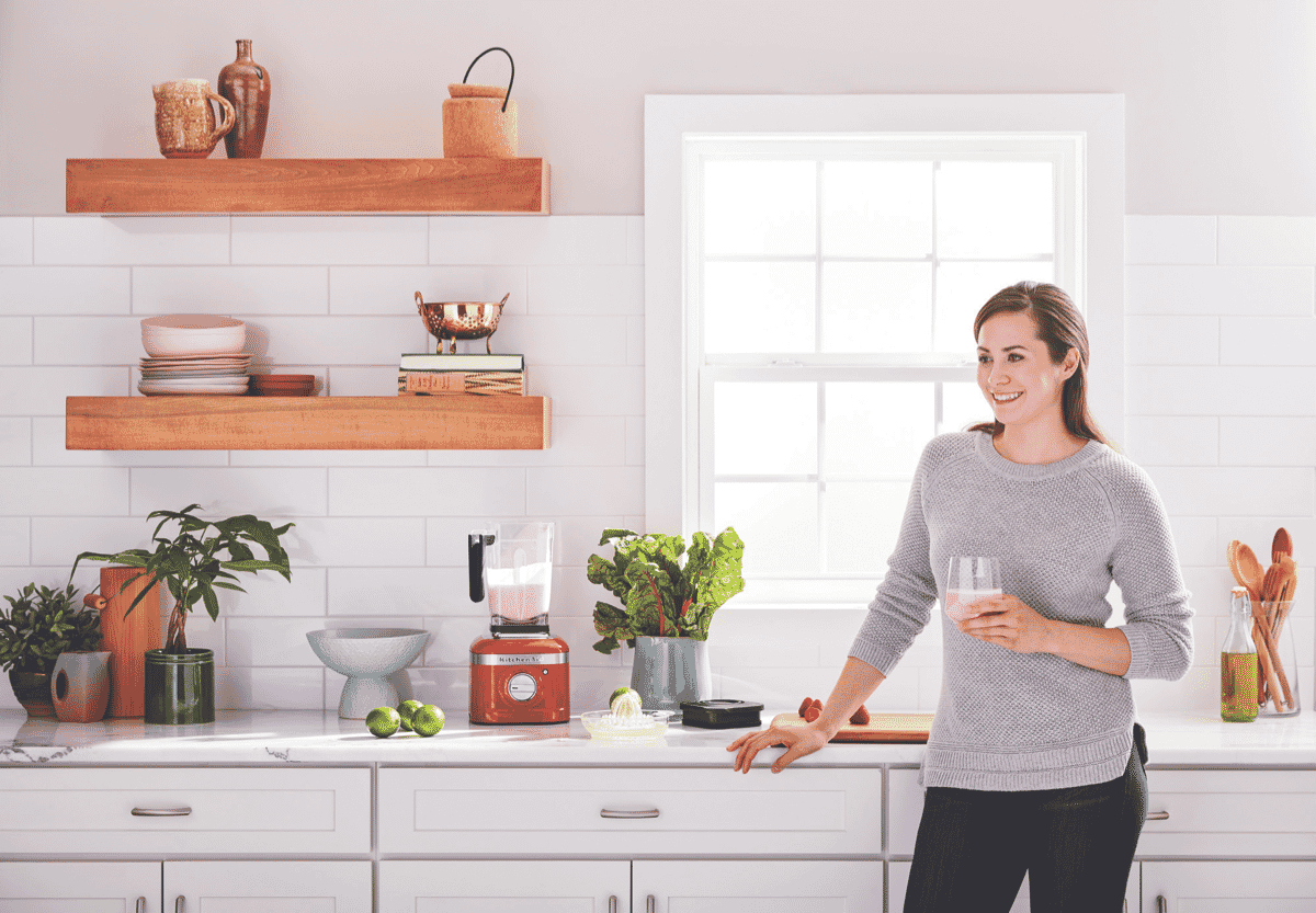 A red KitchenAid Blender sits on a benchtop and a women holds a glass of pink smoothie