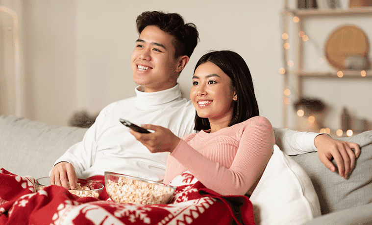 A man and woman watch a movie with popcorn in a living room decorated for Christmas