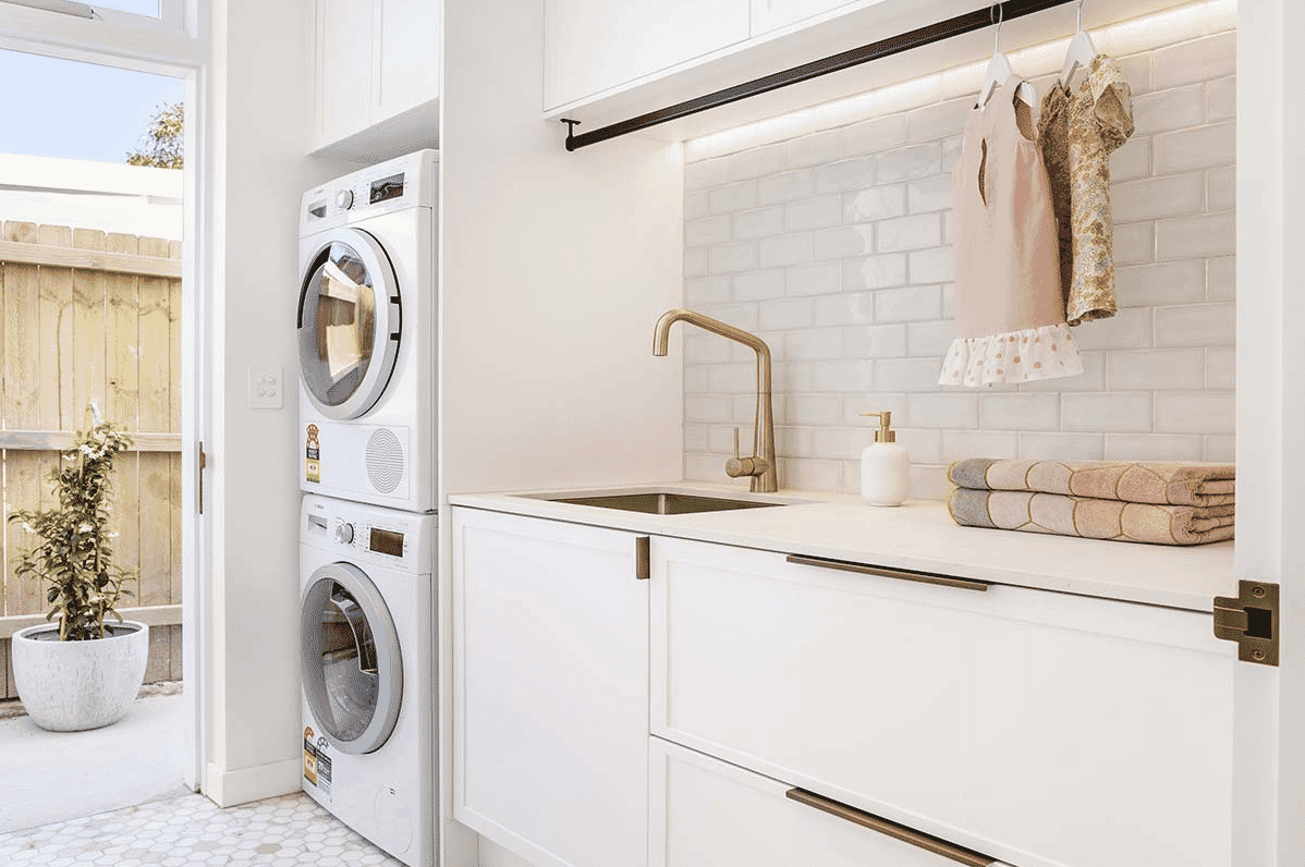 Bright sunny laundry with white cabinetry white subway tiled splashback brass tapware and stacked washing machine and dryer