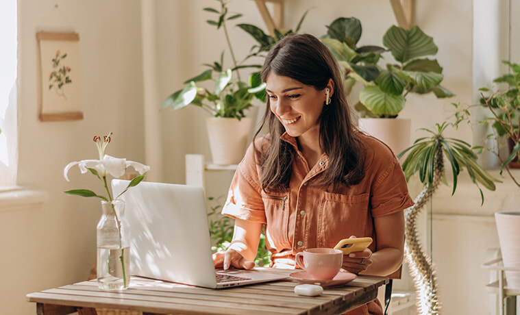 Young woman using a laptop and smartphone while seated in her plantfilled home office