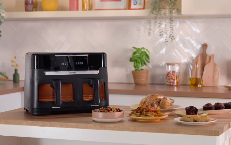 Tefal dual drawer air fryer with viewing windows on a kitchen bench, surrounded by plates of roast chicken, chips, vegetables and muffins.