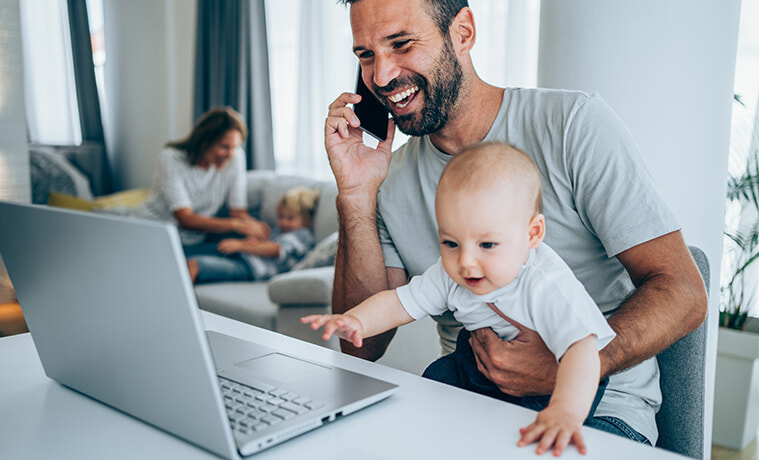 Young handsome father with a baby on his lap using a laptop and mobile phone at home 